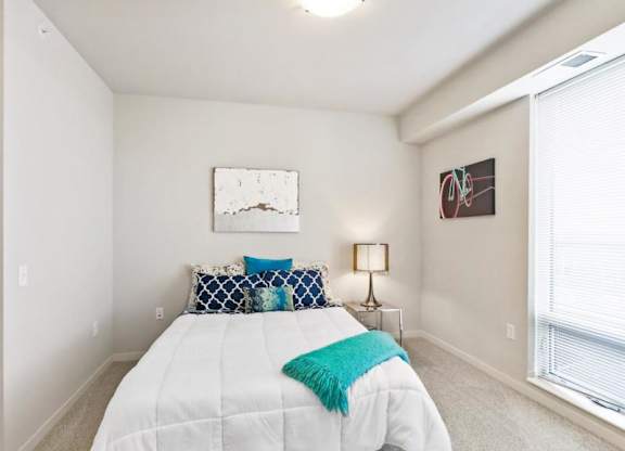 A bright bedroom here at Blue and Lime Apartments with a white bed, teal and navy pillows, abstract wall art, a side lamp, and a large window. The space feels modern and inviting.