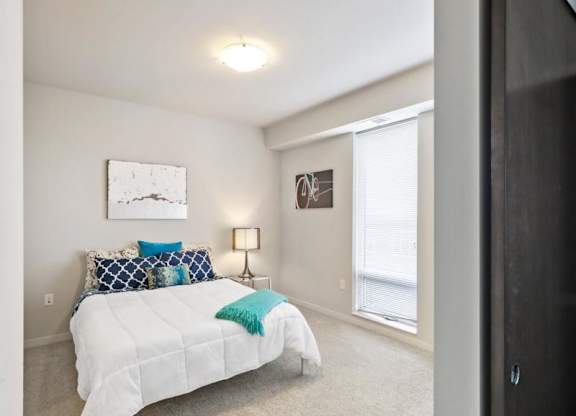 A modern bedroom here at Blue and Lime Apartments with a neatly made bed featuring blue and white pillows, teal accents, abstract art, a lamp, and a large window with blinds.