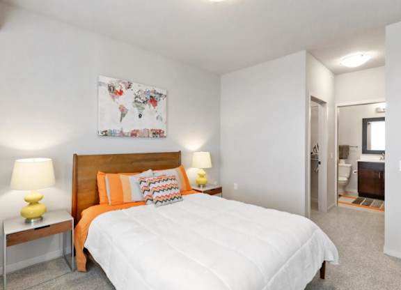 Modern bedroom here at Blue and Lime Apartments with a wooden bed, white and orange bedding, and colorful pillows. Lamps on both sides, world map art above, open view to a bathroom.