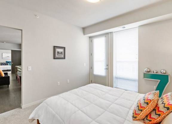 Bright, minimalist bedroom here at Blue and Lime Apartments with white decor, a neatly made bed with colorful cushions, wall art, and sliding glass doors leading to a sunlit balcony.