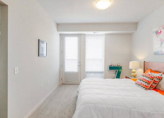 Bright bedroom here at Blue and Lime Apartments with a white comforter, colorful zigzag pillows, nightstand with lamp, wall art, and large windows, creating a cozy, modern vibe.