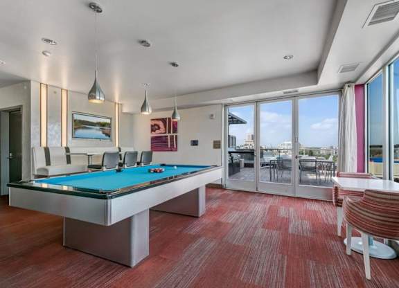 Modern recreation room here at Blue and Lime Apartments with a pool table featuring blue felt, pendant lights, and red carpet. Large windows and patio doors reveal a sunny outdoor view.