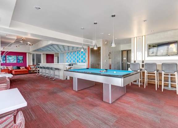 Modern lounge here at Blue and Lime Apartments with a pool table centered on red carpet. Bar stools line a counter to the right, and cozy seating with colorful art is in the back.