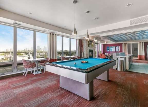 Modern lounge here at Blue and Lime Apartments with a pool table on a red-carpeted floor, large windows showing a city view, and seating areas with red chairs, creating a relaxed vibe.