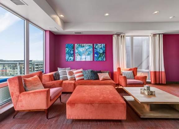 A bright seating area here at Blue and Lime Apartments featuring orange mid-century modern furniture, pink walls, large windows, abstract art, and decorative pillows, conveying a vibrant and cozy atmosphere.