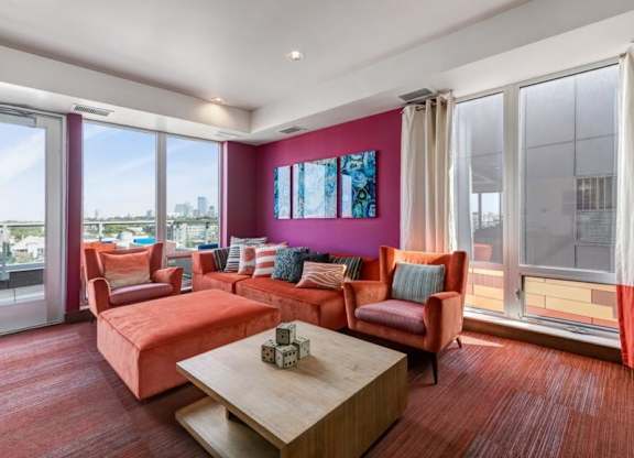 Contemporary seating area here at Blue and Lime Apartments with a pink accent wall, bright orange sofas, colorful cushions, abstract art, and large windows overlooking a cityscape.