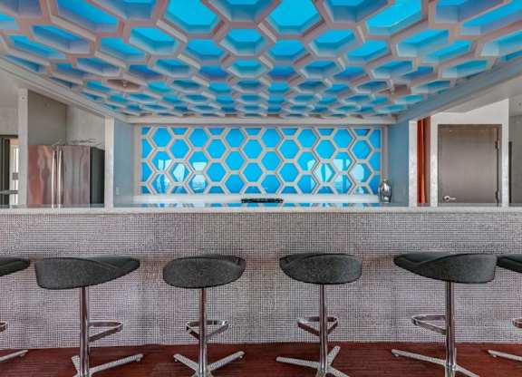 Modern bar interior here at Blue and Lime Apartments with hexagonal blue accents on wall and ceiling, five sleek black stools lined up at a mosaic-tiled counter, creating a stylish ambiance.