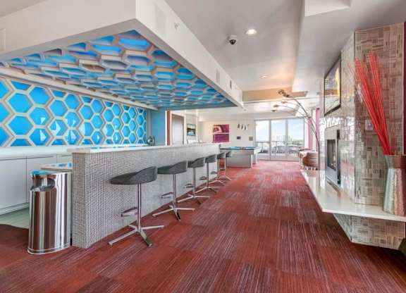 Modern lounge here at Blue and Lime Apartments with red carpet, featuring a bright blue hexagonal wall design. Barstools line a sleek counter. Large windows offer an airy, inviting feel.