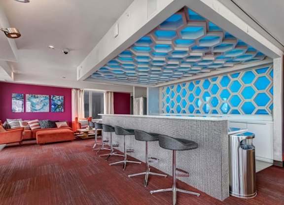 Modern lounge here at Blue and Lime Apartments with a stylish bar, hexagonal blue ceiling, and a row of barstools. Bright orange sofas by wall art create a vibrant, cozy atmosphere.