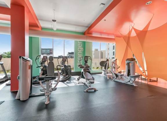 Bright, modern gym here at Blue and Lime Apartments with colorful orange and green walls. Features stationary bikes and exercise machines by large windows, creating an inviting atmosphere.