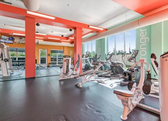 Bright, modern gym here at Blue and Lime Apartments with large windows, treadmills, and elliptical machines. Red beams and colorful walls create an energetic atmosphere.