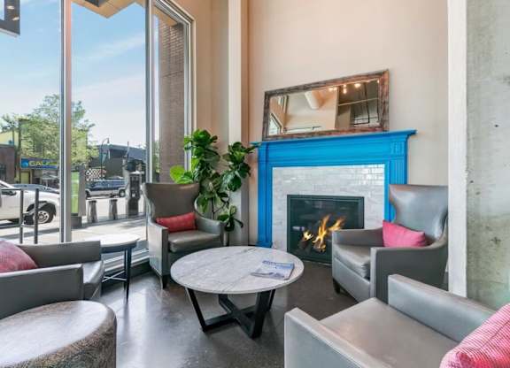 Cozy lobby corner here at Blue and Lime Apartments with a lit fireplace framed in blue, surrounded by modern gray chairs with red cushions. Large windows offer outdoor views.