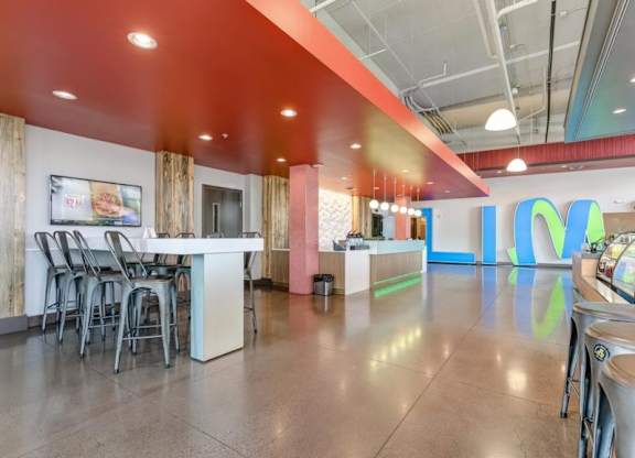 Modern brightly lit lobby here at Blue and Lime Apartments with high ceilings and polished floors. Features high tables, metal chairs, an orange ceiling, and colorful wall letters "LIME."