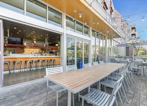 Outdoor patio here at Blue and Lime Apartments with long wooden table, surrounded by metal chairs. Inside, a modern bar with stools and plants. String lights create a welcoming ambiance.