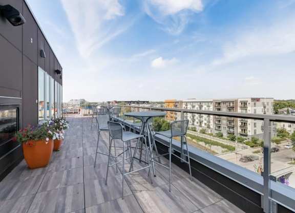 Rooftop patio here at Blue and Lime Apartments with tall tables and chairs overlooking city buildings. Clear blue sky, potted flowers on wooden deck, modern and relaxed ambiance.