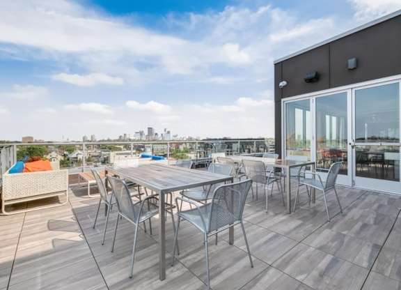 Rooftop patio here at Blue and Lime Apartments with metal tables and chairs, overlooking city skyline. Bright sky enhances an inviting, relaxed atmosphere. Large glass doors nearby.