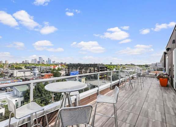 Rooftop patio here at Blue and Lime Apartments with metal tables and chairs overlooks a cityscape under a vibrant blue sky with clouds. Potted plants add color to the modern scene.