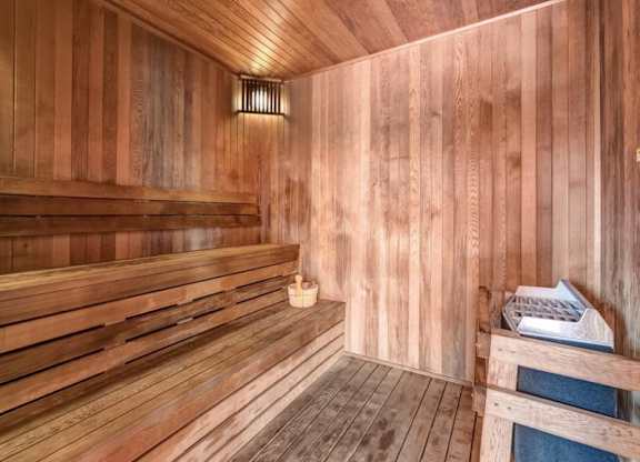 Wooden sauna interior here at Blue and Lime Apartments with warm lighting, featuring tiered benches on the left, a wall-mounted light above, and a heater with stones and a wooden bucket on the right.