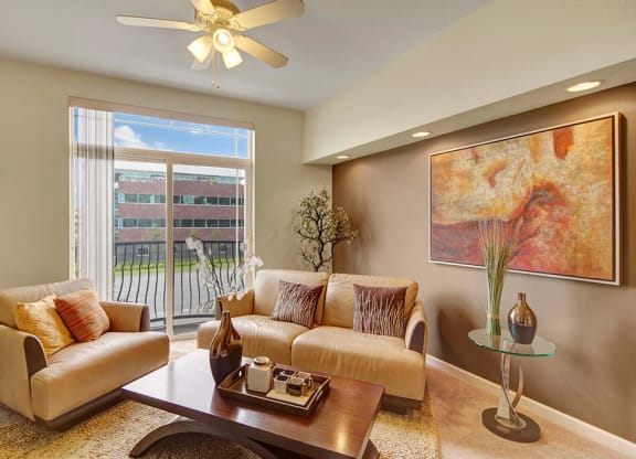 Bright modern living room here at Park Plaza 2 Apartments with beige sofas, abstract painting, ceiling fan, and large window showing a view of an office building. Cozy and inviting.