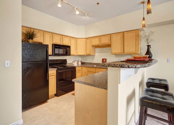 Modern kitchen here at Park Plaza 2 Apartments with light wood cabinets and black appliances, including a fridge and stove. Granite countertops, pendant lighting, and bar stools add elegance.