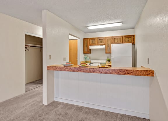 Compact kitchen here at Park Plaza Apartments with a wood-accented breakfast bar, set with colorful plates. White appliances, wood cabinets, and an open closet create a cozy, inviting feel.
