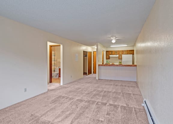 Spacious empty living room here at Park Plaza Apartments with beige carpet and cream walls, leading to a kitchen with brown cabinets and a bathroom visible on the left. Cozy and inviting.