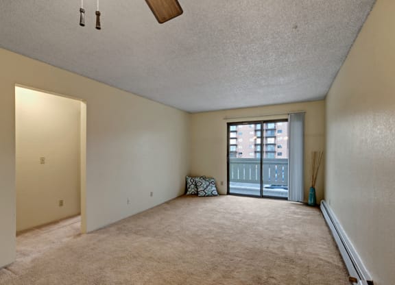 Spacious, empty living room here at Park Plaza Apartments with beige walls and carpet, ceiling fan, and sliding glass door opening to a balcony. Minimalist decor includes a cushion and a vase.