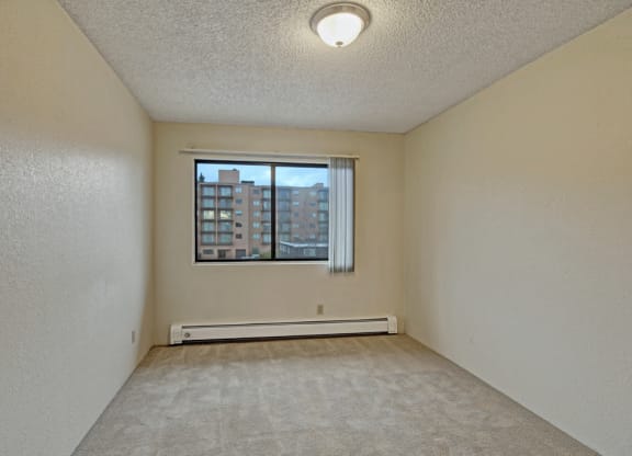 Bedroom here at Park Plaza Apartments with beige walls and carpet is lit by a ceiling fixture. A window with vertical blinds reveals a view of nearby buildings, creating a calm atmosphere.