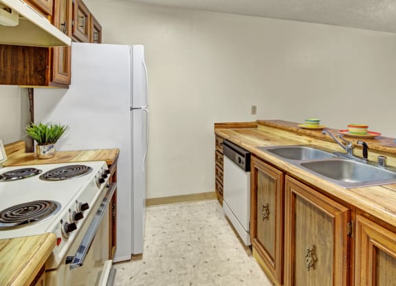 Cozy kitchen here at Park Plaza Apartments with wood cabinets, beige countertop, double sink, white appliances, and multicolored bowls atop the counter, exuding a vintage charm.