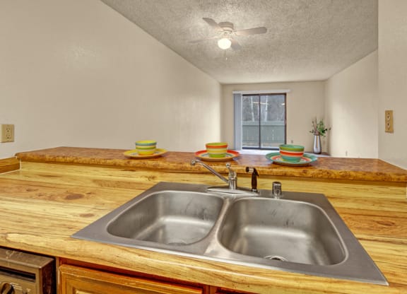Cozy kitchen here at Park Plaza Apartments with a wooden countertop and double sink, set with colorful plates and bowls. A ceiling fan and distant sliding doors enhance the room's brightness.