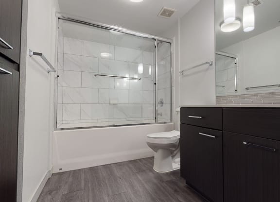 Modern bathroom here at Escape Apartments with dark wood cabinets, a large mirror, and a double sink with chrome faucets. The bathtub has a glass door and white tile walls.