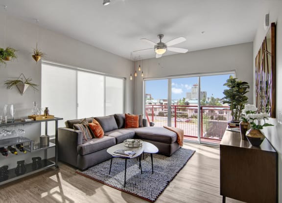 Modern living room here at Proxy 333 Apartments with a gray sectional sofa, colorful cushions, and geometric wall decor. Large windows overlook a sunny cityscape. Cozy and bright ambiance.