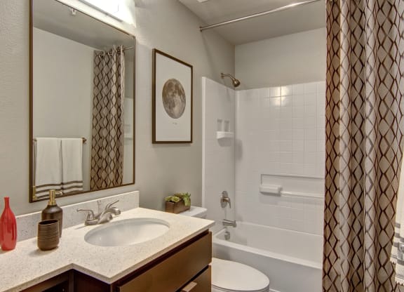 Modern bathroom here at Proxy 333 Apartments with a granite countertop and oval sink. A red and brown vase sits beside a soap dispenser. A framed moon print, patterned shower curtain, and white towels add a contemporary touch.