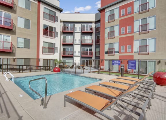 Modern Proxy 333 Apartment courtyard with a small, rectangular pool surrounded by orange lounge chairs and red accents. Balcony views and a clear, sunny sky.