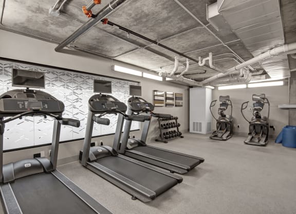 Modern gym here at Proxy 333 Apartments with gray treadmills and elliptical machines on a light carpet. Dumbbells are neatly stacked on a rack, and geometric wall design adds style.