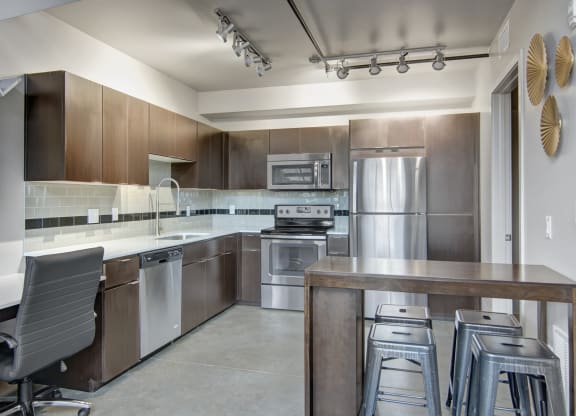 Modern kitchen here at Proxy 333 Apartments with dark wood cabinets, stainless steel appliances, and a sleek countertop. A high table with metal stools stands nearby. Warm, neutral tones.