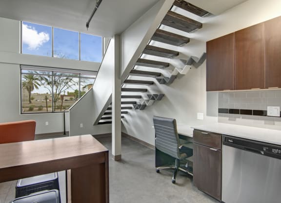 Modern interior here at Proxy 333 Apartments with a floating staircase, large windows showing a palm tree and blue sky. Features a small office area with dark cabinets. Calm atmosphere.