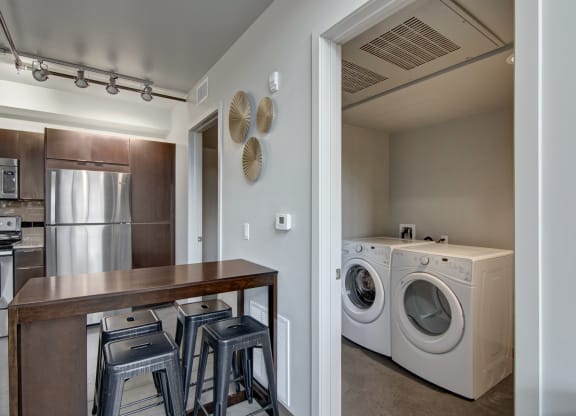 Modern kitchen here at Proxy 333 Apartments with wood cabinets, stainless steel appliances, and barstool seating. Adjacent laundry room with washer and dryer, minimalist decor.