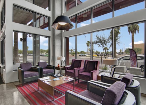 Modern lobby here at Proxy 333 Apartments with large windows, purple chairs around a wooden coffee table on a red-striped rug. Bright lighting, outside view of palm trees and parked cars.