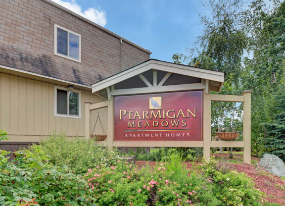 Sign reading "Ptarmigan Meadows Apartment Homes," surrounded by lush greenery and flowers, sits in front of a beige building under a partly cloudy sky.