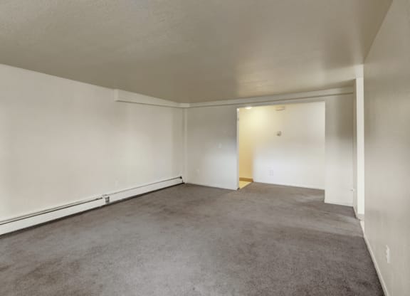 Empty living room here at Ptarmigan Meadows with beige carpet and off-white walls. Open doorway leads to a small, lighted area. Sparse, calm atmosphere with a minimalist feel.