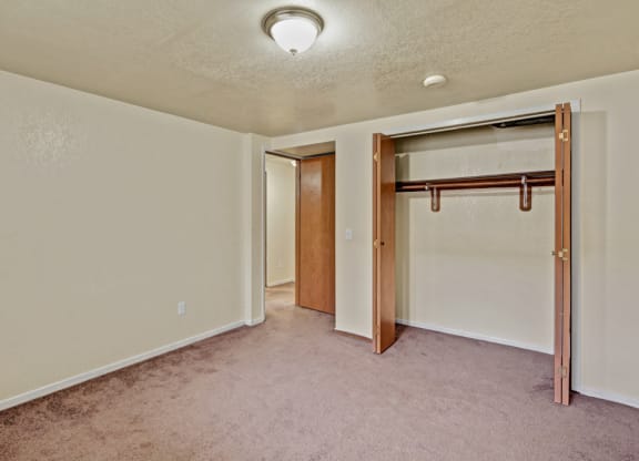 An empty bedroom here at Ptarmigan Meadows with beige walls and a brown carpet. Open closet with wooden doors and a hanging rod. Ceiling light illuminates the space, creating a simple, neutral tone.