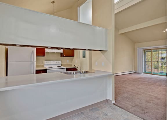 Open-plan kitchen and living room here at Ptarmigan Meadows with neutral tones. Kitchen features white appliances and brown cabinets. Living area has large windows and balcony access.