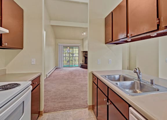 Narrow kitchen here at Ptarmigan Meadows with dark wood cabinets, double stainless steel sink, and white stove. Leads to a carpeted living room with large glass doors.