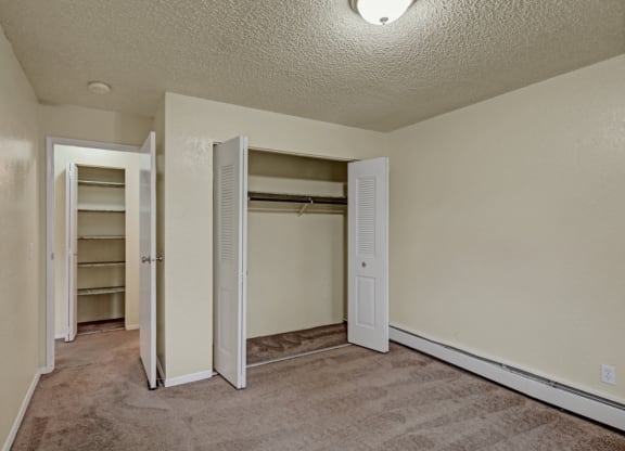 Small empty bedroom here at Ptarmigan Meadows with beige walls and carpet, featuring an open closet with louvered doors and a doorway leading to a shelf-lined closet, lit by a ceiling light.