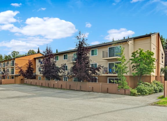 Three-story beige Quiet Creek Apartment building with brown trim, surrounded by trees and enclosed by a wooden fence, under a bright blue sky with scattered clouds.