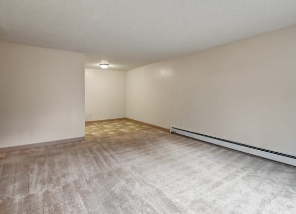 Empty living room here at Quiet Creek Apartments with beige carpet flooring, light cream walls, and a baseboard heater on the right. A lit ceiling light is visible in the back corner. Minimal and clean atmosphere.