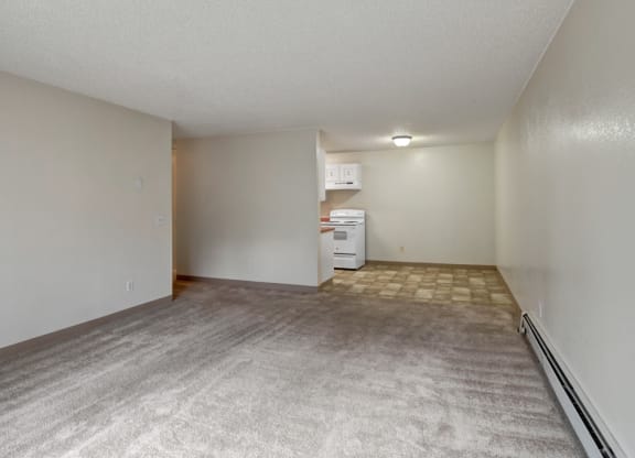 Empty apartment here at Quiet Creek Apartments with beige carpeted living area, leading to a small kitchen with white appliances and linoleum floor. Bright, clean, and minimalist.