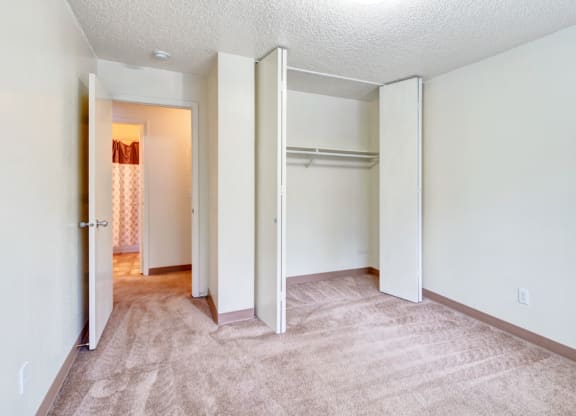 Empty bedroom here at Quiet Creek Apartments with beige carpet and white walls, featuring an open closet with shelves. A door leads to a lit hallway with a patterned shower curtain.