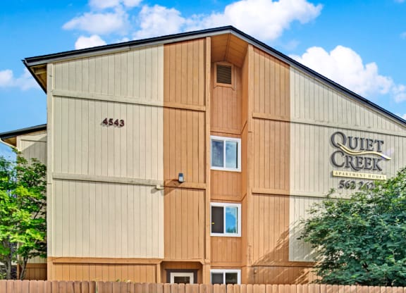 Apartment building with wood paneling in beige and tan. "Quiet Creek Apartment Homes" sign on the right. Trees and a wooden fence in the foreground. Blue sky above.